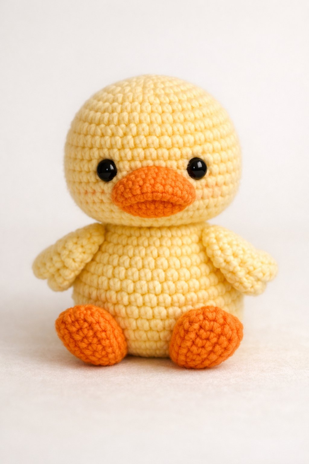 A handmade crocheted yellow duck with orange beak and feet sitting upright on a plain background.